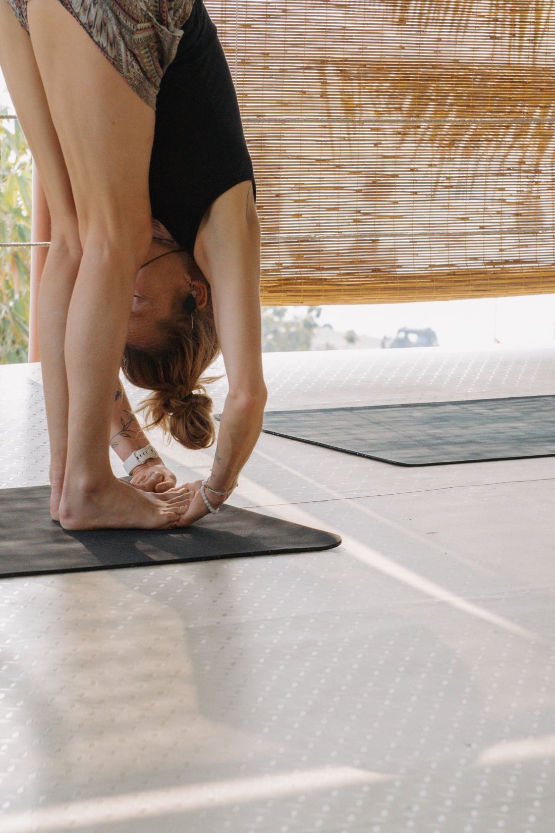 A standing forward fold on the shala mat beside bamboo blinds